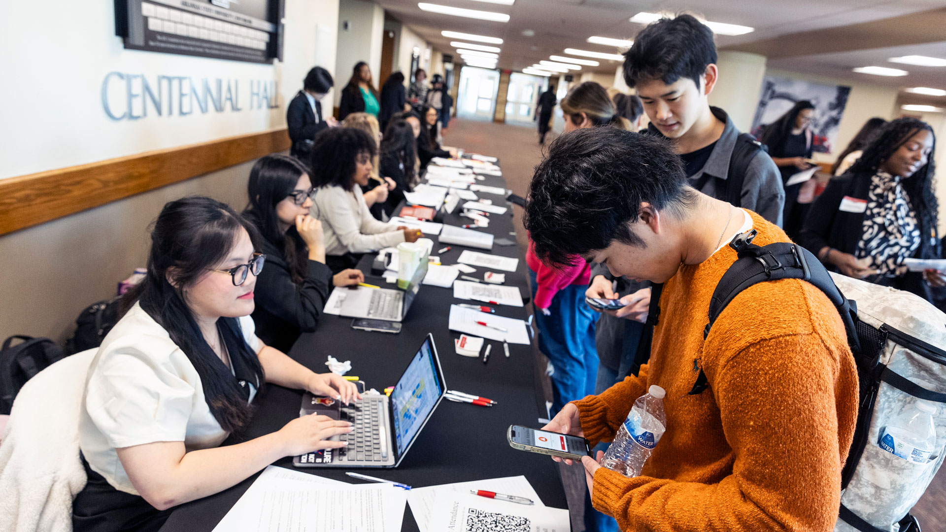 several workers with laptop computers are seated behind a table. Students on the other side of the table are getting information, using their phones to scan QR codes.