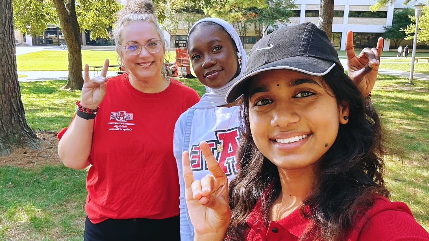 3 women are smiling and posing for the camera outdoors on a sunny day. They are using their hand to make the 